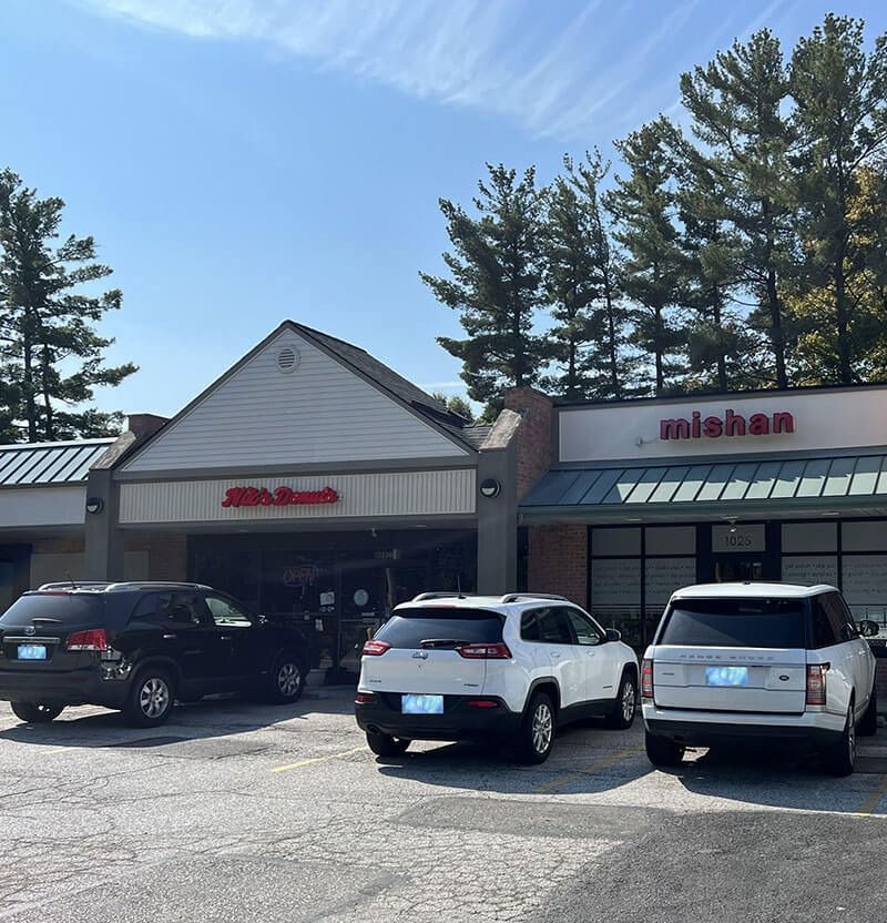 Exterior view of a strip mall with three parked cars. Stores include 