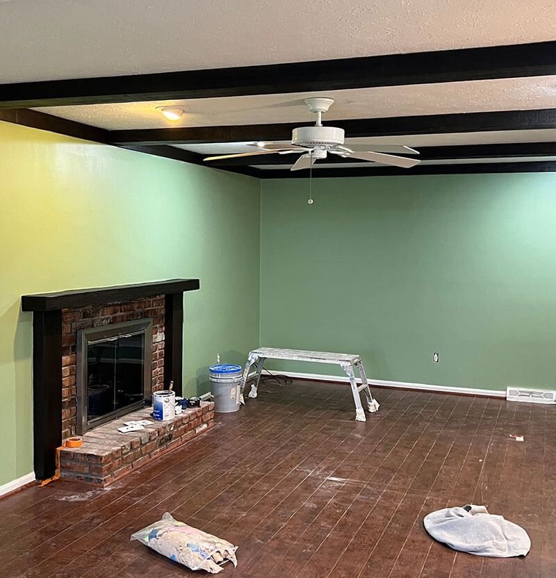 Living room with green walls, dark ceiling beams, fireplace, wooden floor, and a ceiling fan.