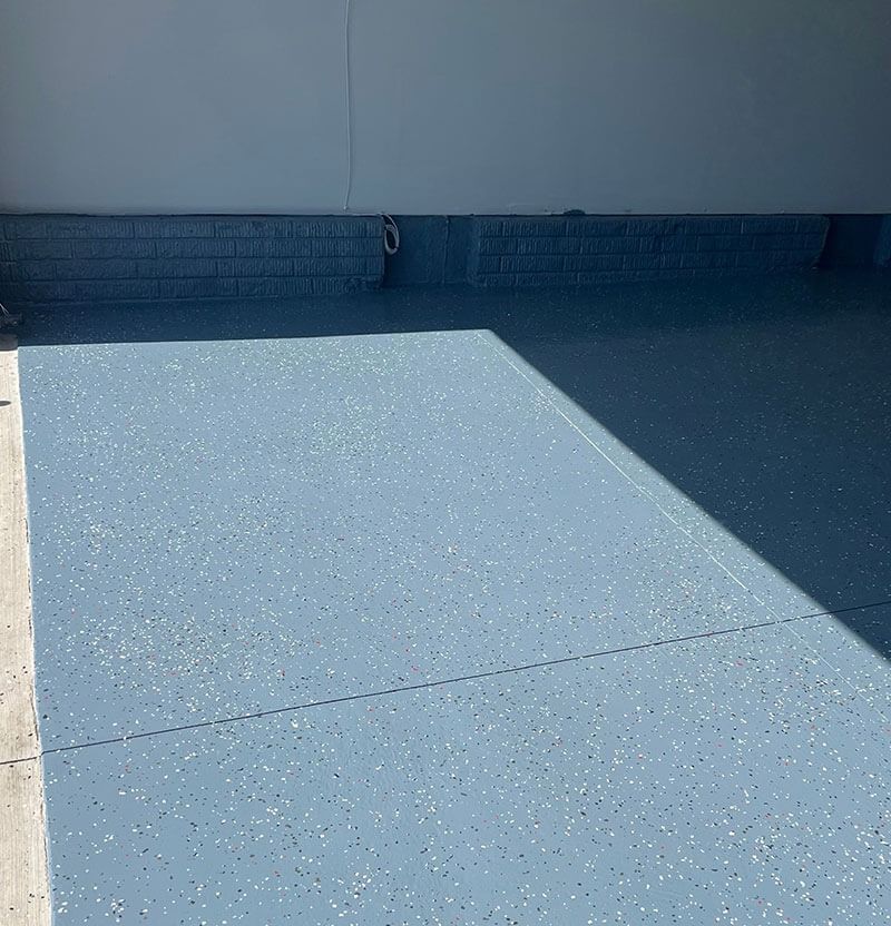 Blue epoxy garage floor with speckled chips; a white wall and brick border.