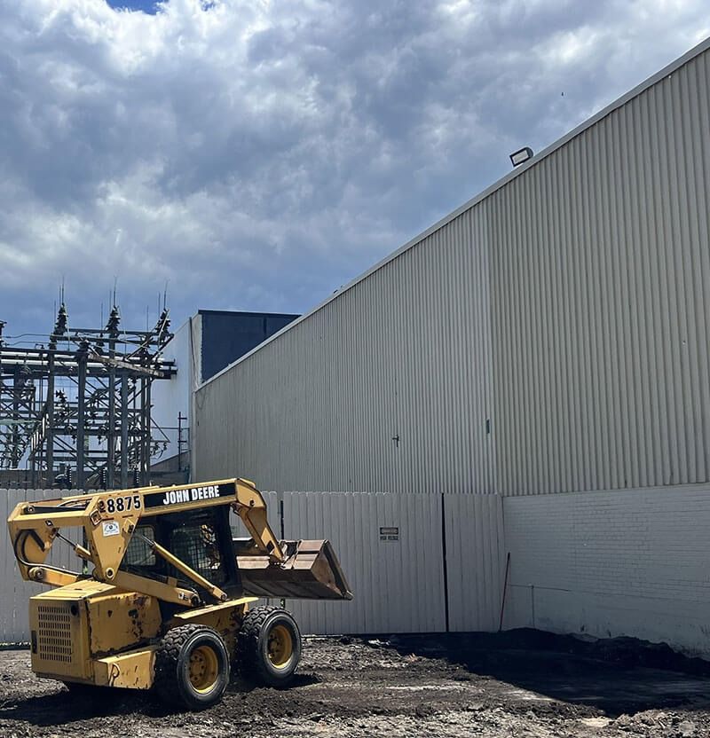 Yellow skid steer loader near a white industrial building and power lines under a cloudy sky.