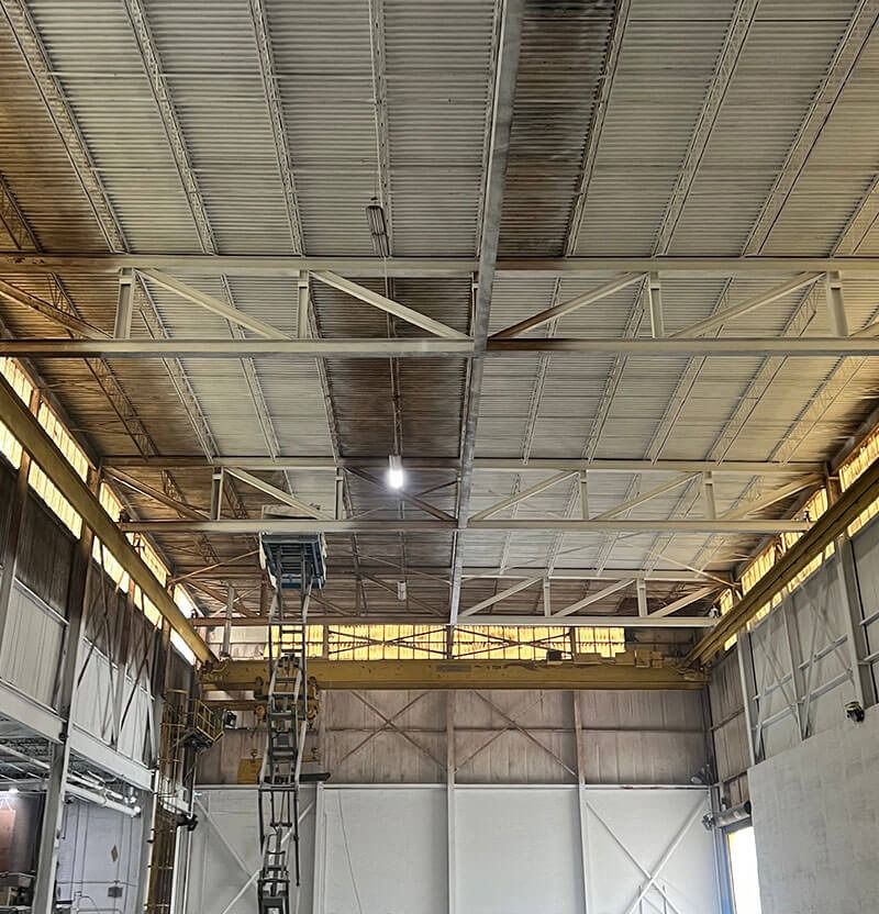 Warehouse interior with metal roof and beams, stained walls, and industrial equipment.