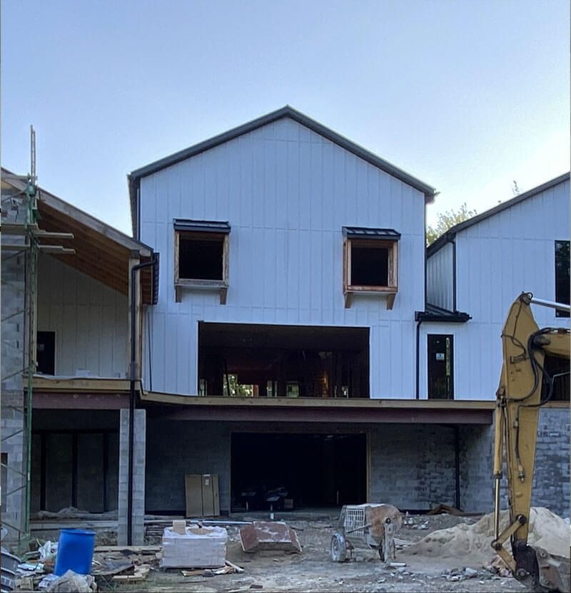 Construction site of a two-story building with white siding and exposed steel beams; excavator in the foreground.