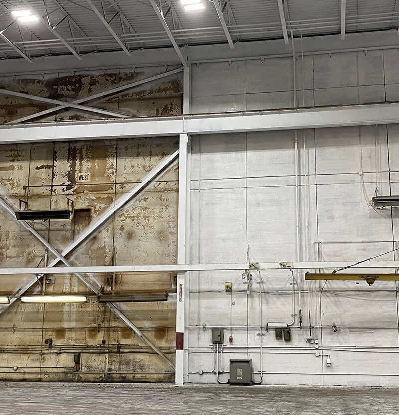 A warehouse interior. Left side shows aged wall; right side, a newly painted white wall with equipment.