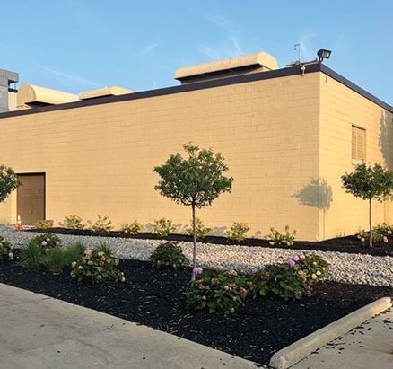Tan brick building with landscaped flower beds, small trees, and a concrete border.