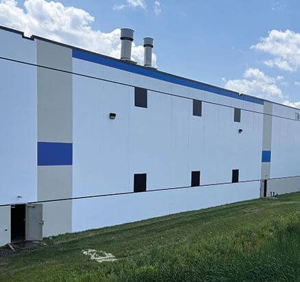 White and blue industrial building with vents on the roof and several dark square windows, set against a blue sky.