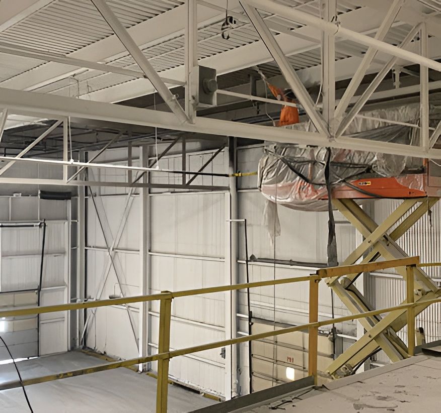 Worker on scissor lift painting white ceiling beams in a large industrial building.