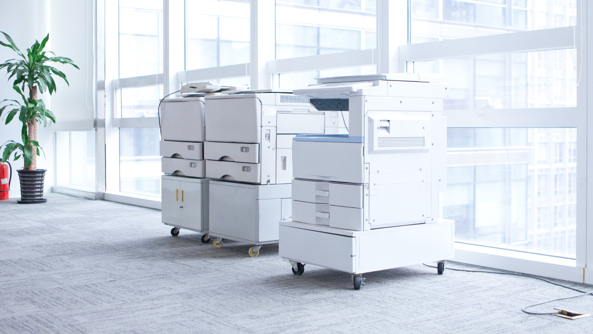 A row of printers are sitting in a room next to a window.