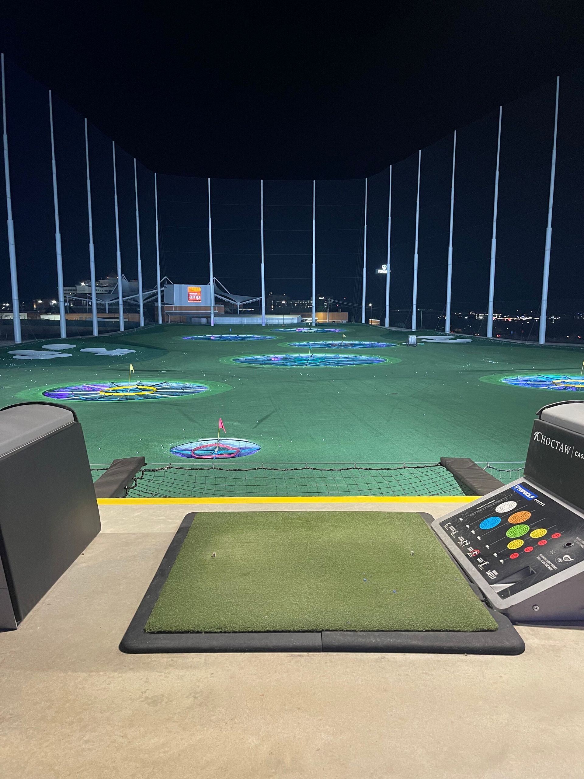Driving range at night with green hitting bays, target flags, and illuminated field lights.