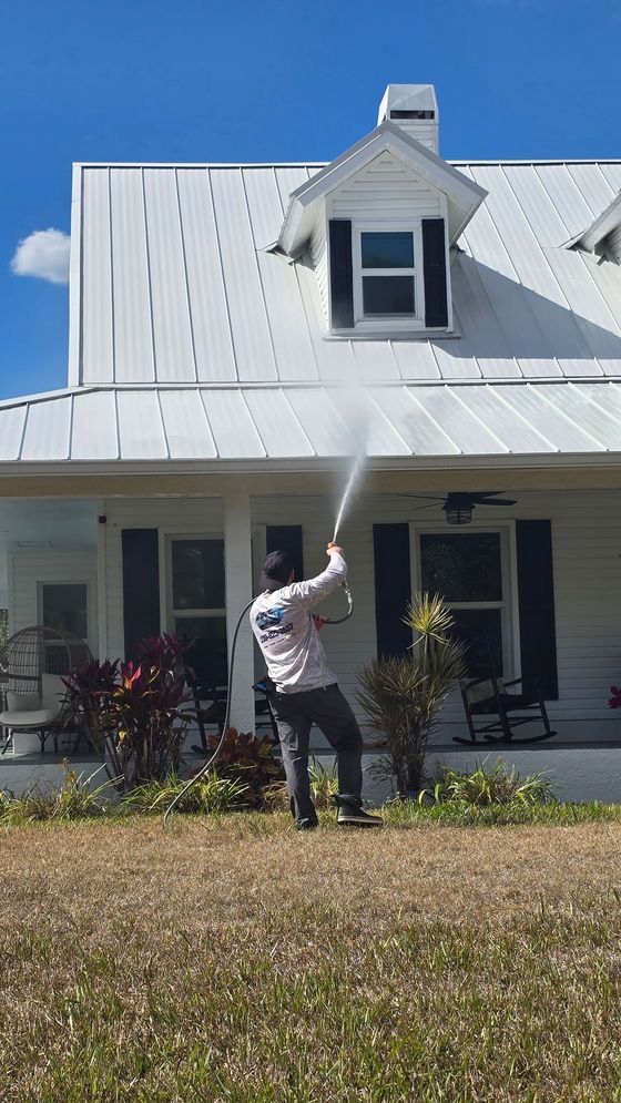 A person in a white long-sleeved shirt pressure washes the white metal roof of a house under a clear blue sky.