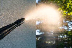 A pressure washer nozzle sprays a fine mist of water onto a textured, light-colored wall outdoors.