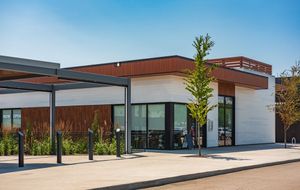 A modern one-story building with white and wood-paneled walls, large glass windows, and a shaded outdoor seating area.