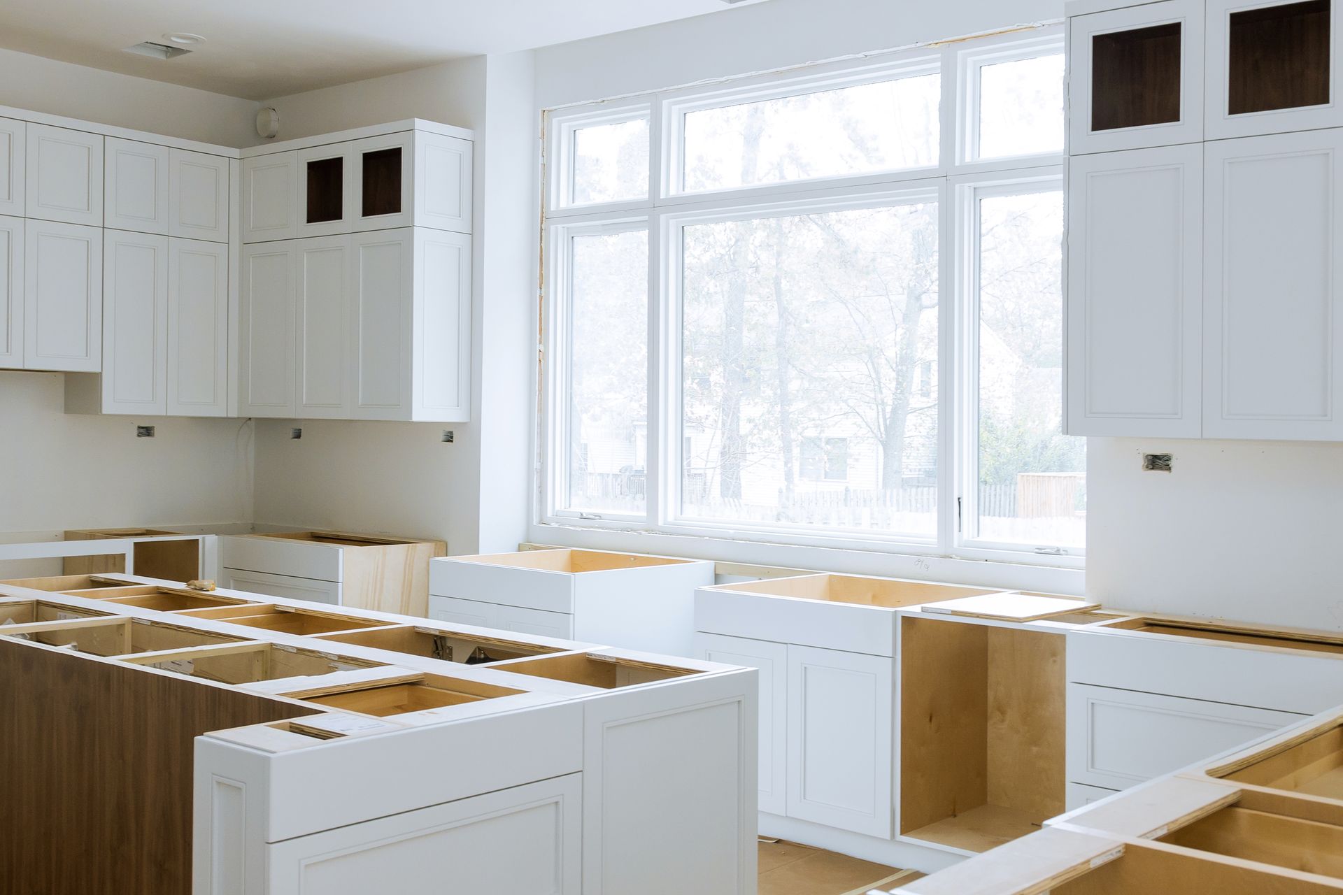 Kitchen cabinets being installed, with white uppers and unfinished wood lowers, by a large window.