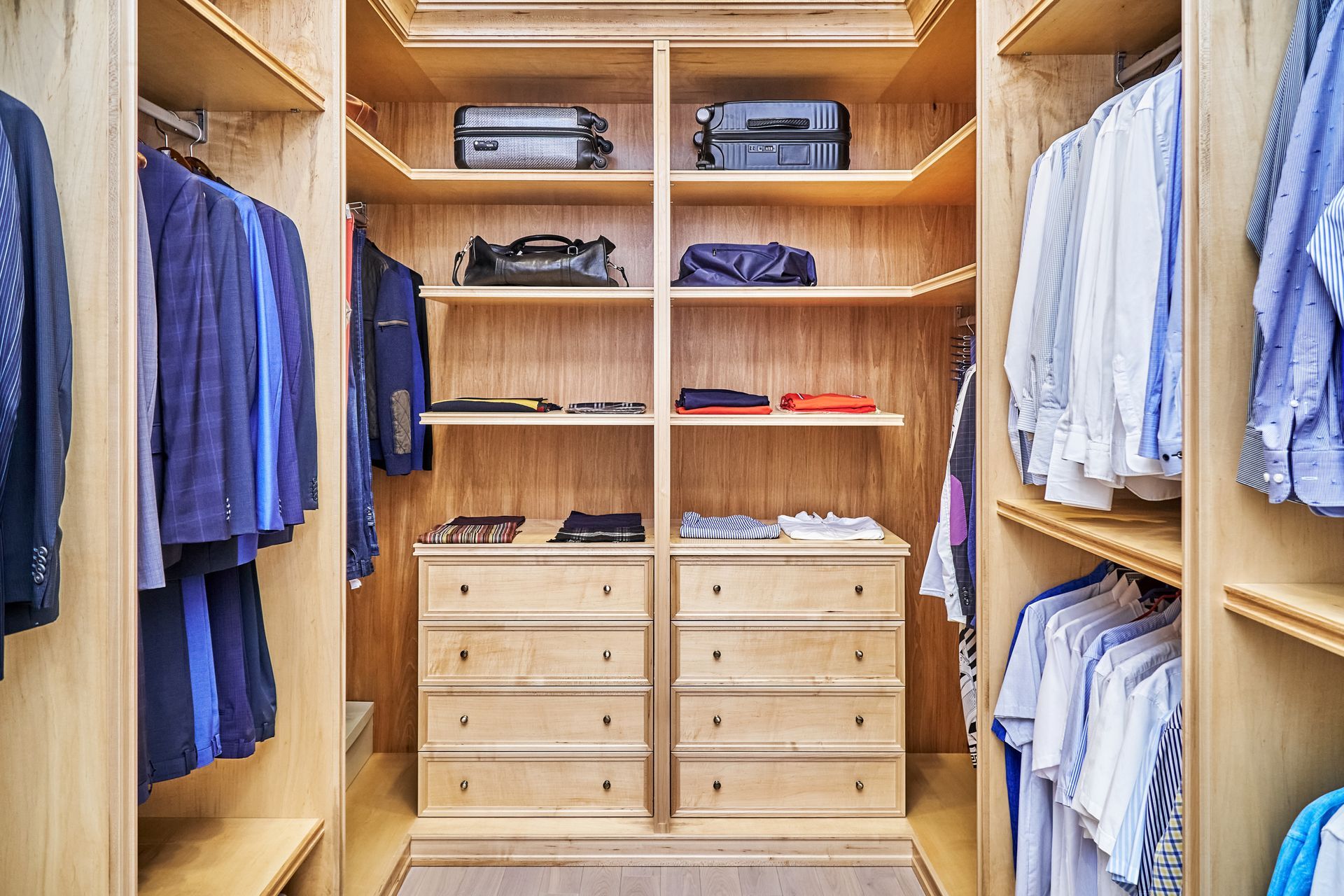 Walk-in closet with hanging clothes, shelves holding bags, and drawers, all made of light wood.