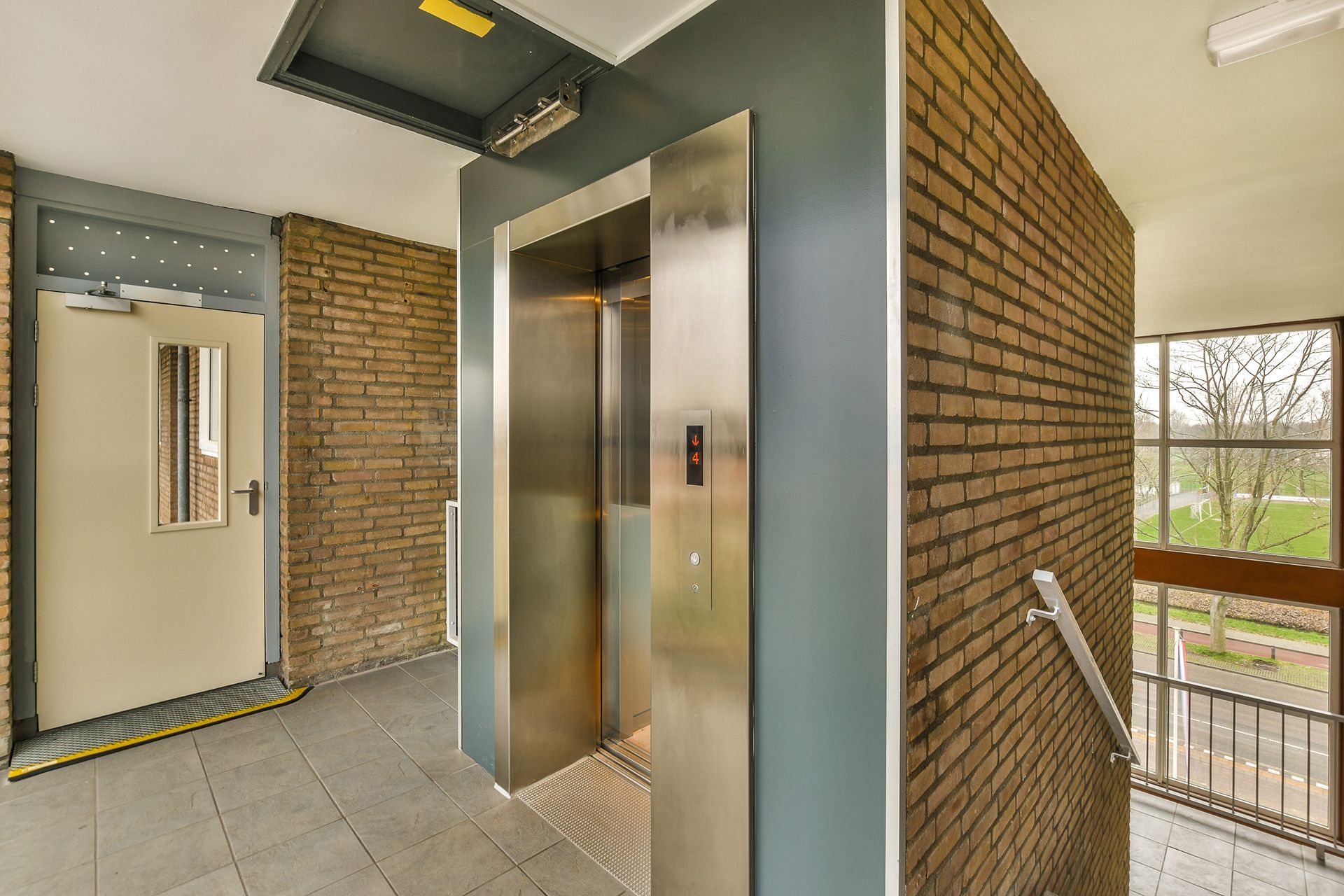 Ascensor con puertas abiertas de acero inoxidable en un pasillo de ladrillo junto a una escalera con vistas desde una ventana.