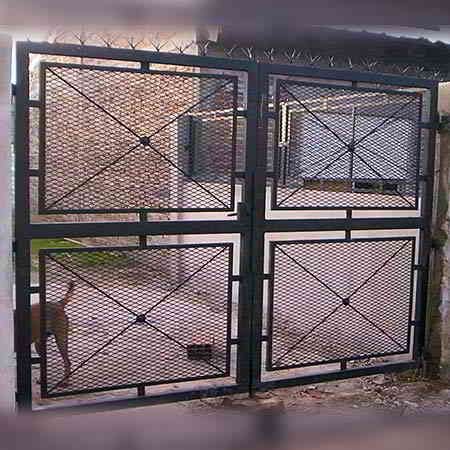 Puerta metálica negra con paneles de malla romboidal; se ve un perro detrás de una de las puertas.