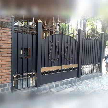 Puerta metálica negra con elementos decorativos, pilar de ladrillo y acera.