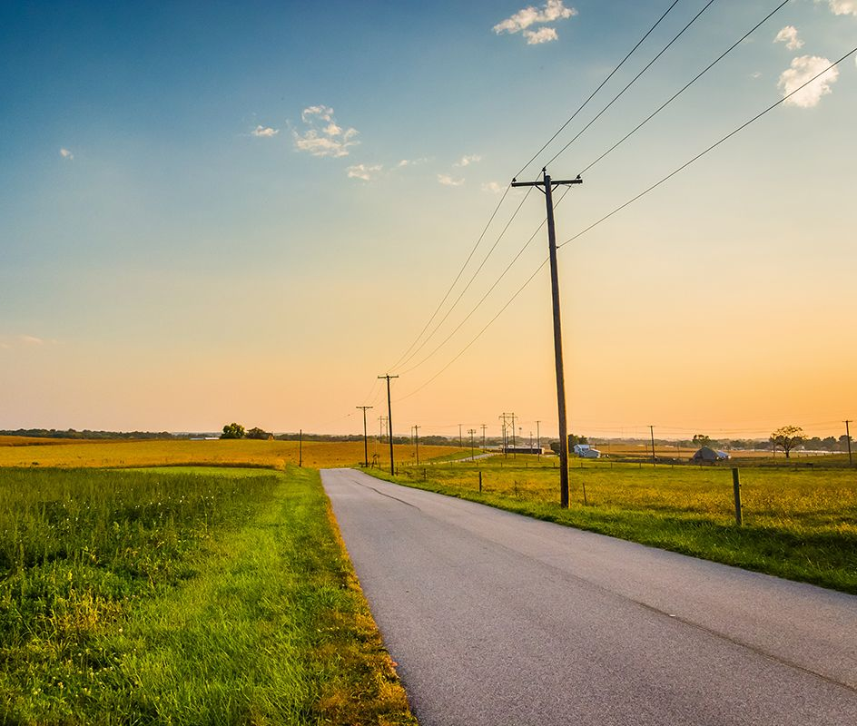 Road through fields with telephone poles under a colorful sunset.