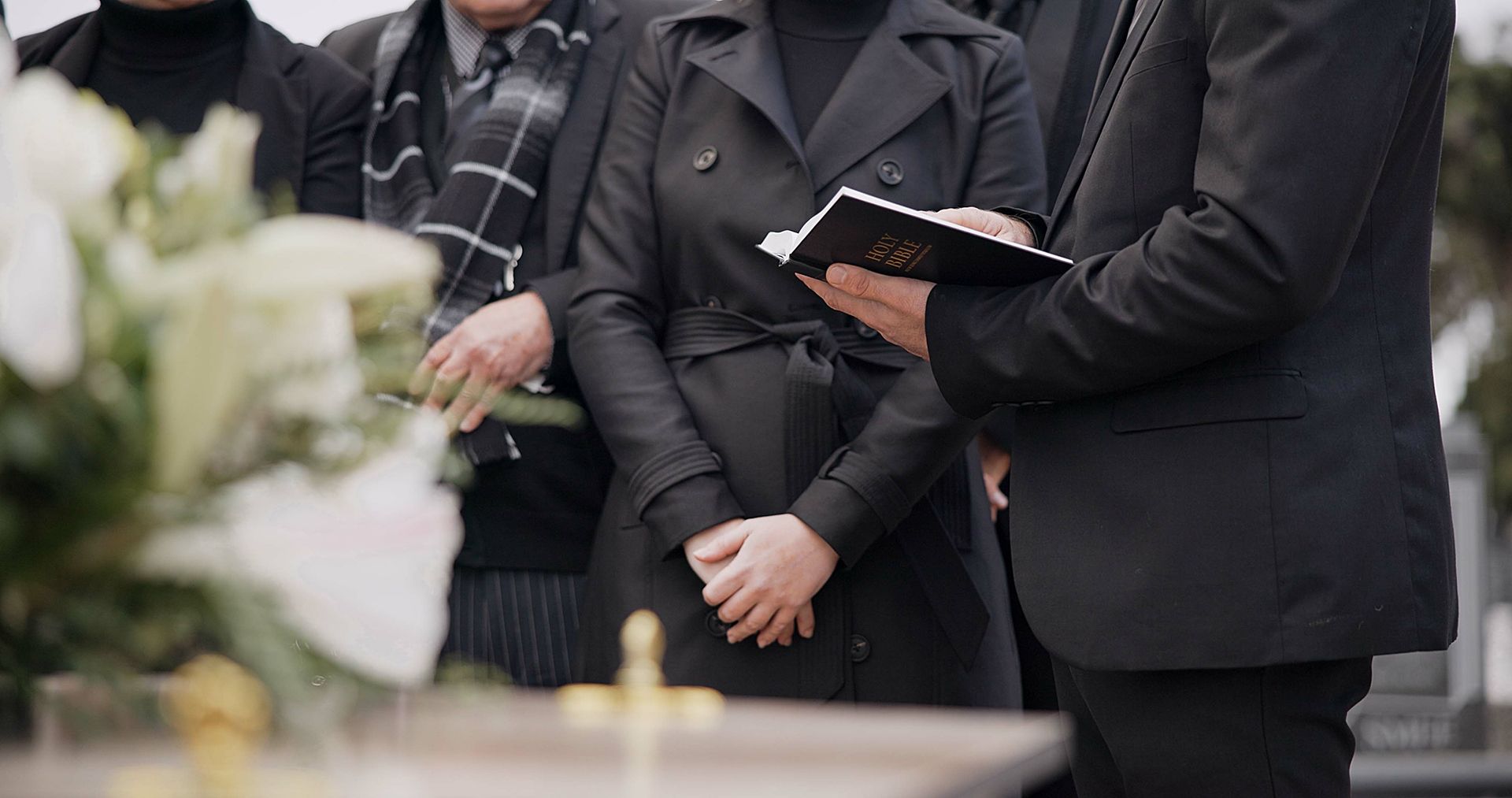 People in black attire at a funeral service, standing near a coffin and flowers.