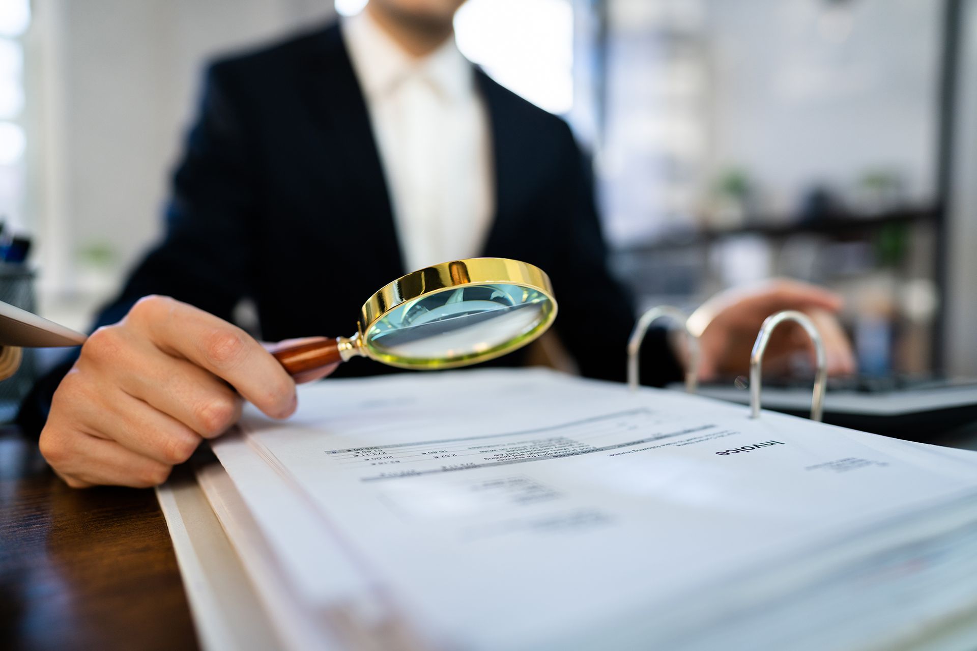 Person in suit using magnifying glass to inspect documents in an office setting.