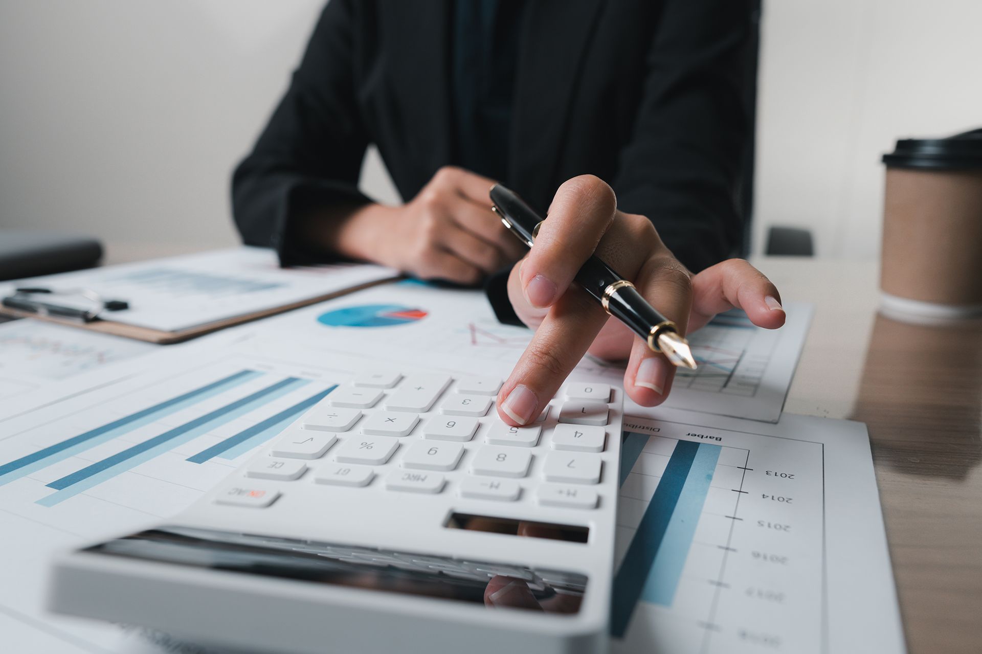 Person in a black blazer using a calculator, surrounded by financial documents and a coffee cup.