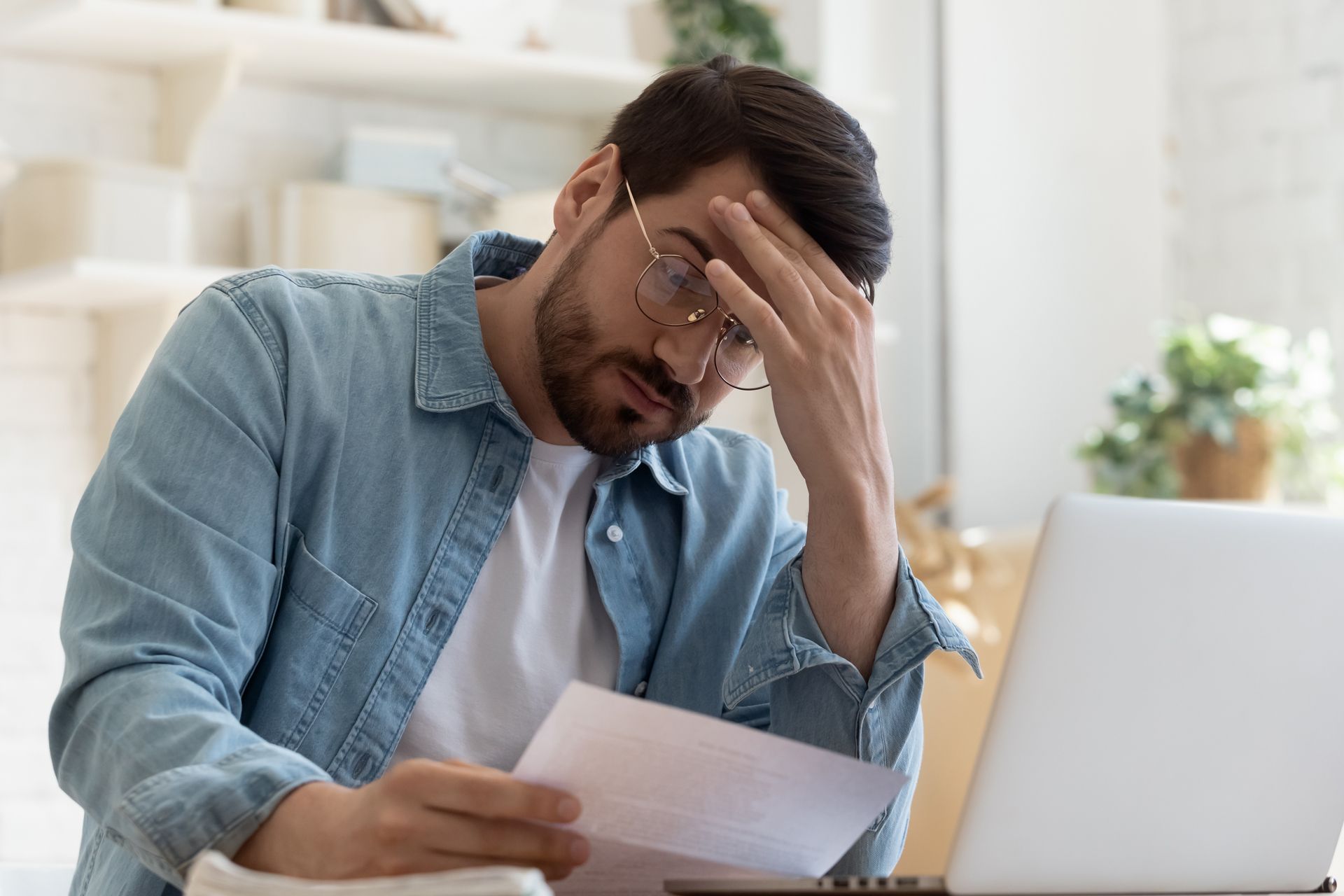 Man with glasses stressed, holding a document, hand on forehead, laptop in front, indoor setting