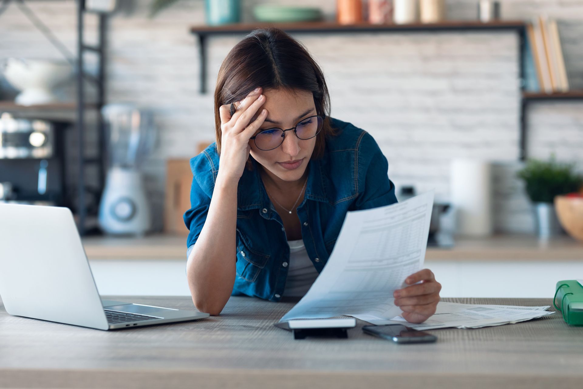 Woman in glasses looks stressed while reviewing documents at a table with a laptop and calculator in a kitchen