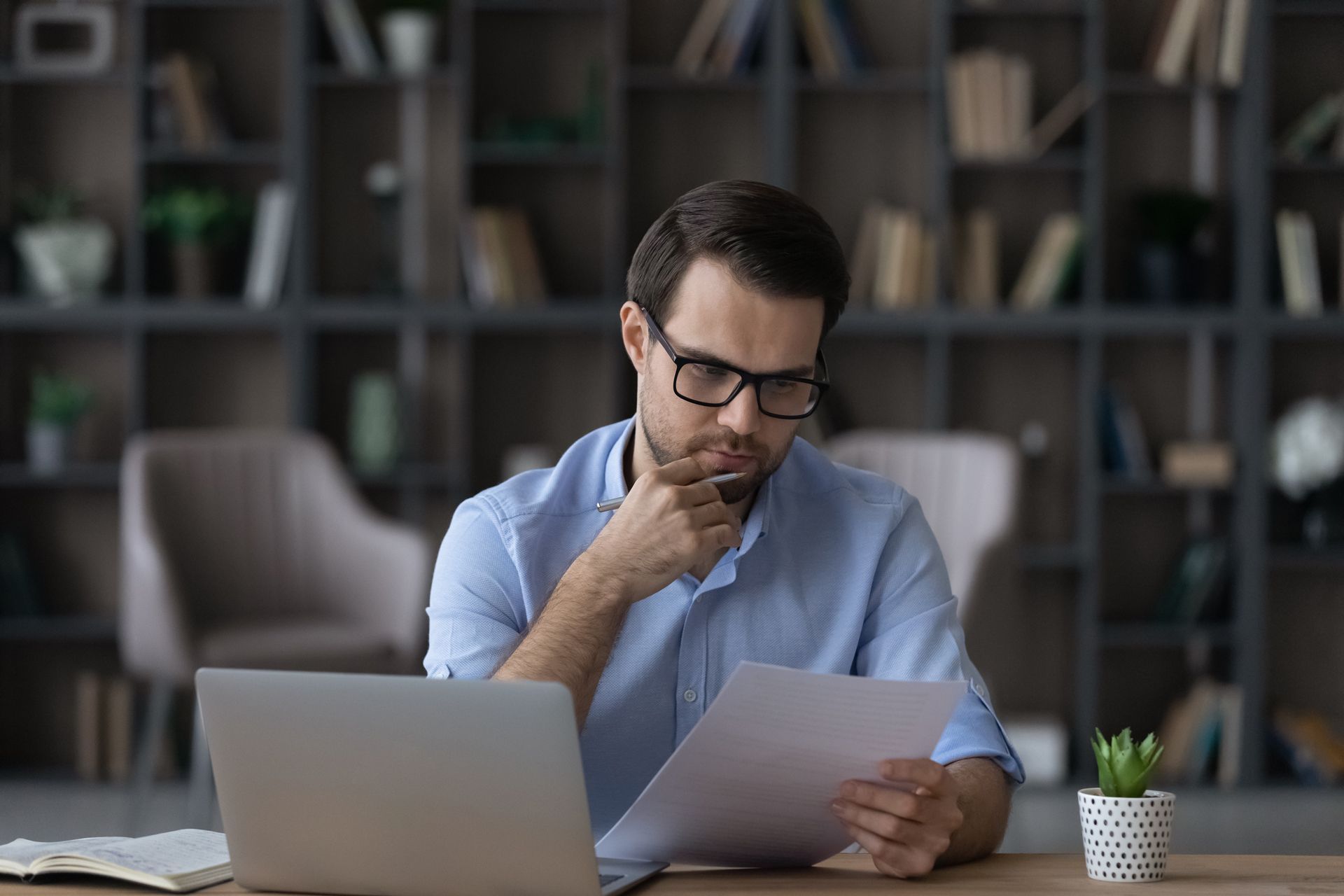 Man in glasses reviews documents at a desk with a laptop; bookshelf background.