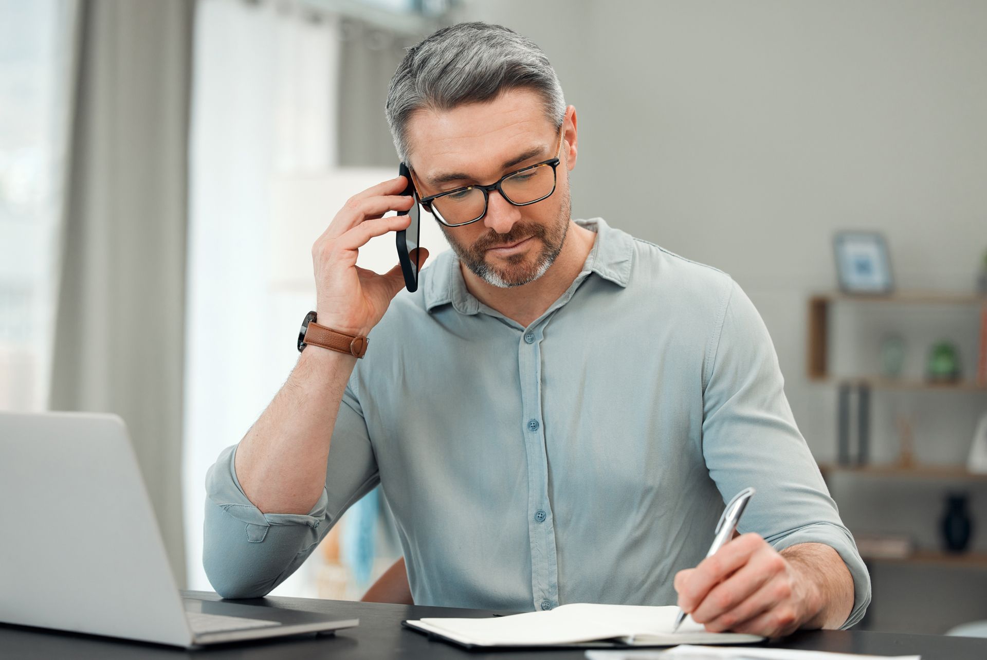 Man on phone, writing in notebook at a desk with a laptop in a home office setting