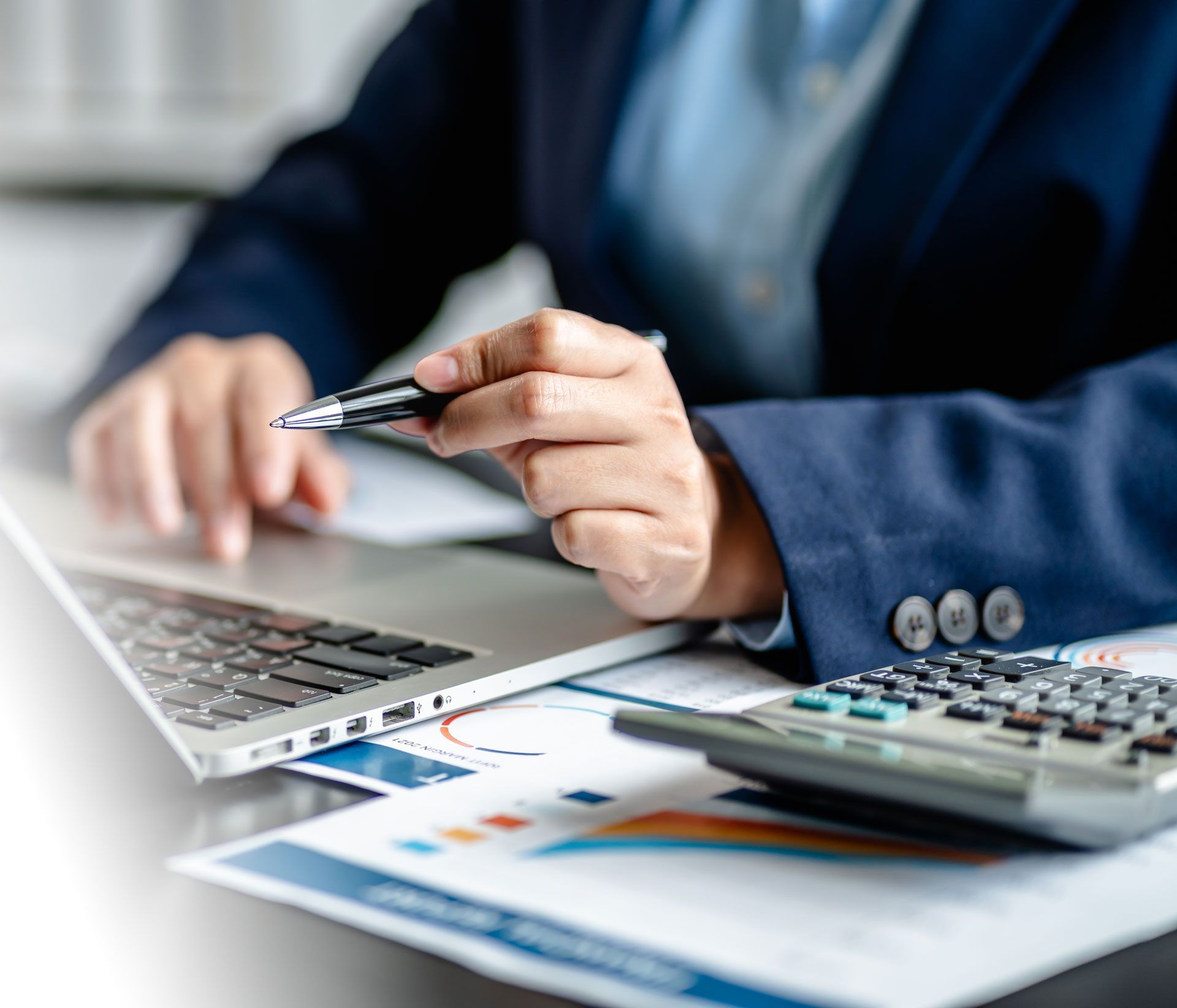 Person using a laptop and calculator beside printed charts, holding a pen in an office setting