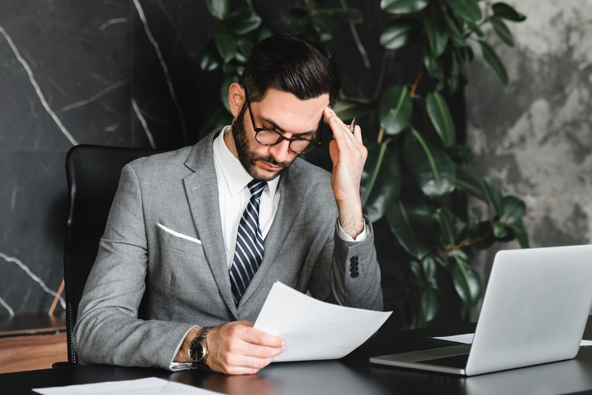 Man in suit, glasses, and tie reviews document, appears stressed, at desk with laptop