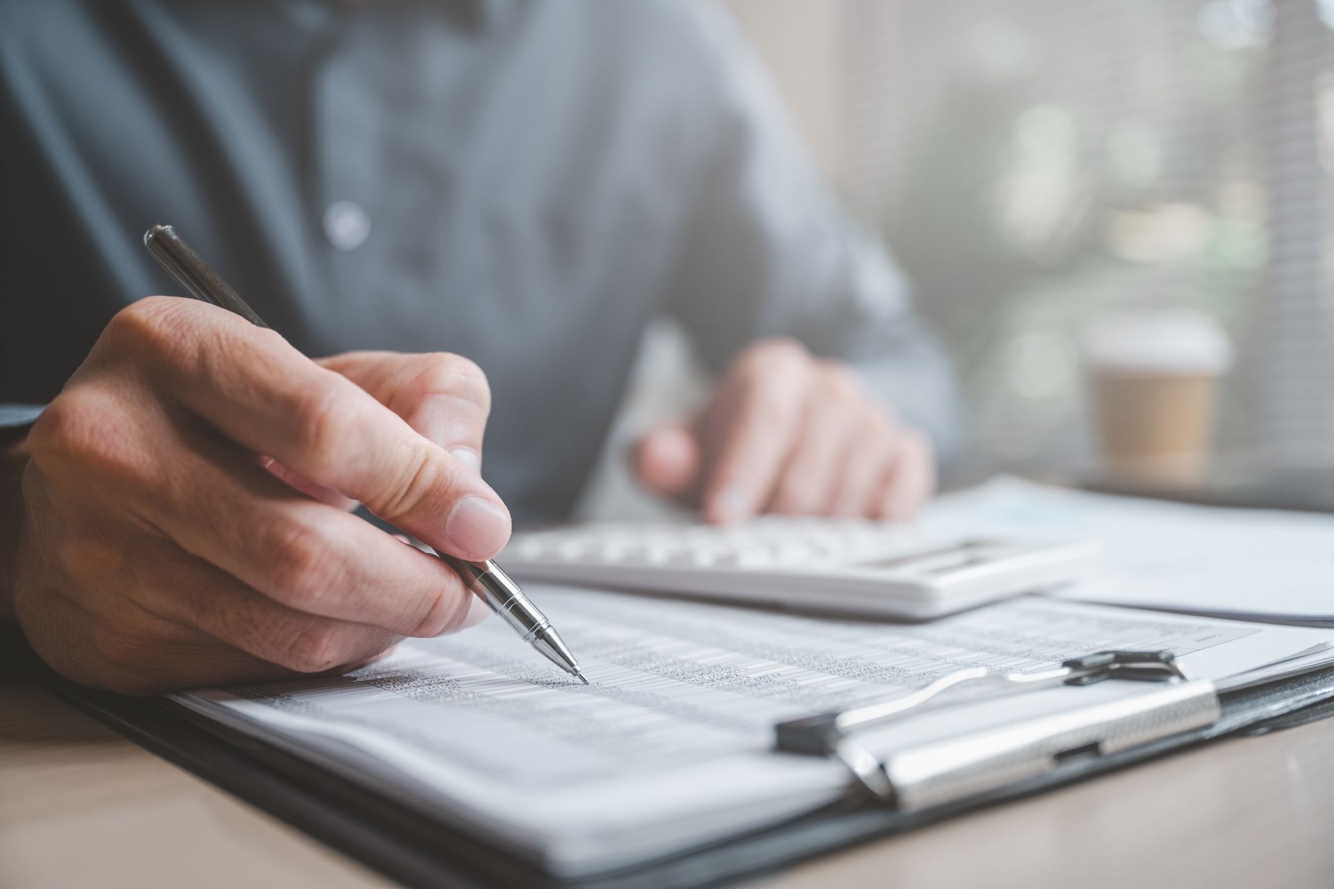 Person writing on a document with a pen, using a calculator; office setting.