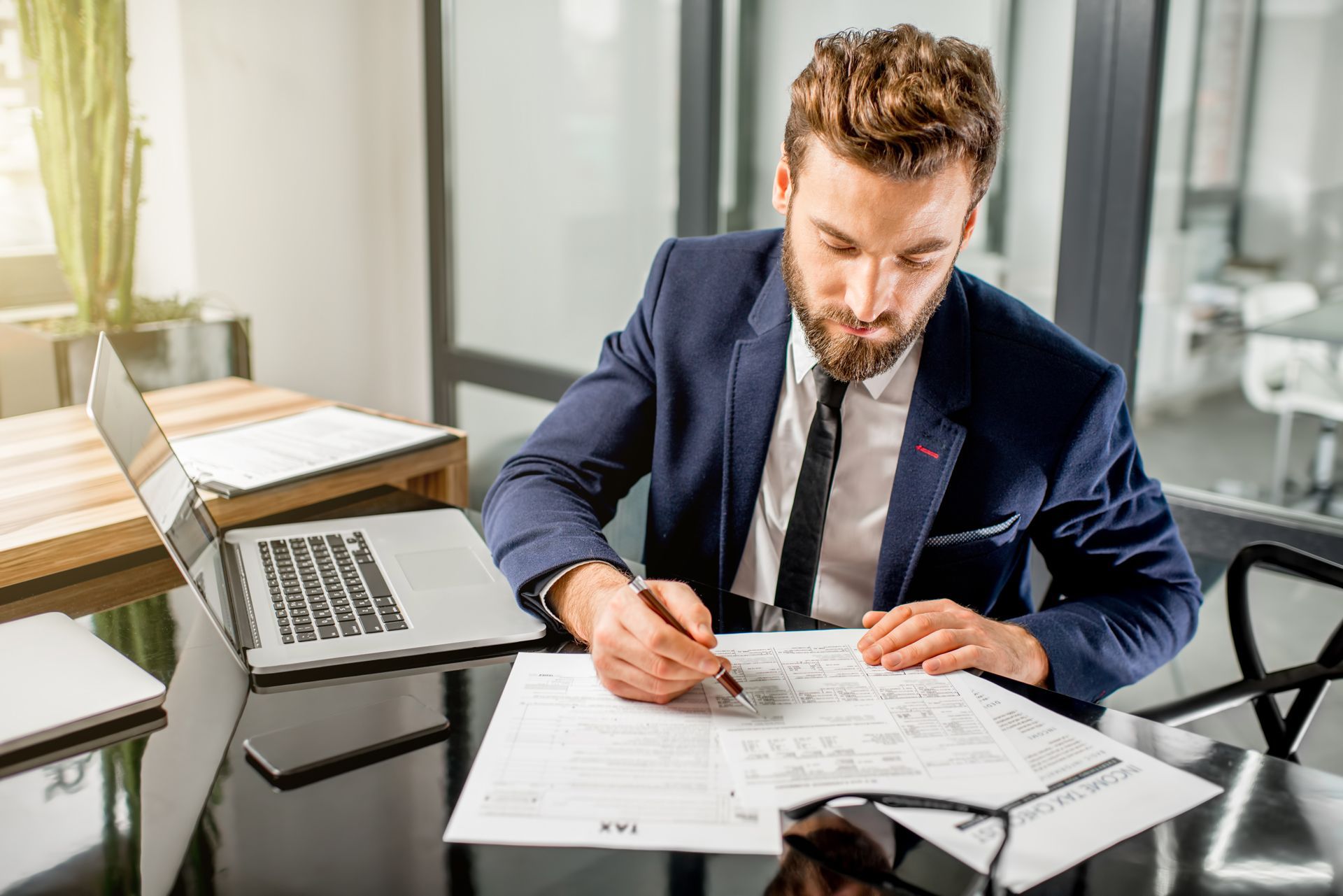 Man in blue suit writing on documents at a desk with a laptop and glasses