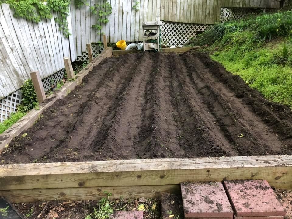 A wooden raised garden bed filled with dirt and bricks in a backyard.