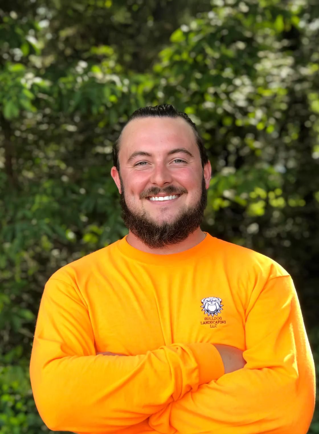 A man with a beard is wearing an orange shirt and smiling with his arms crossed.