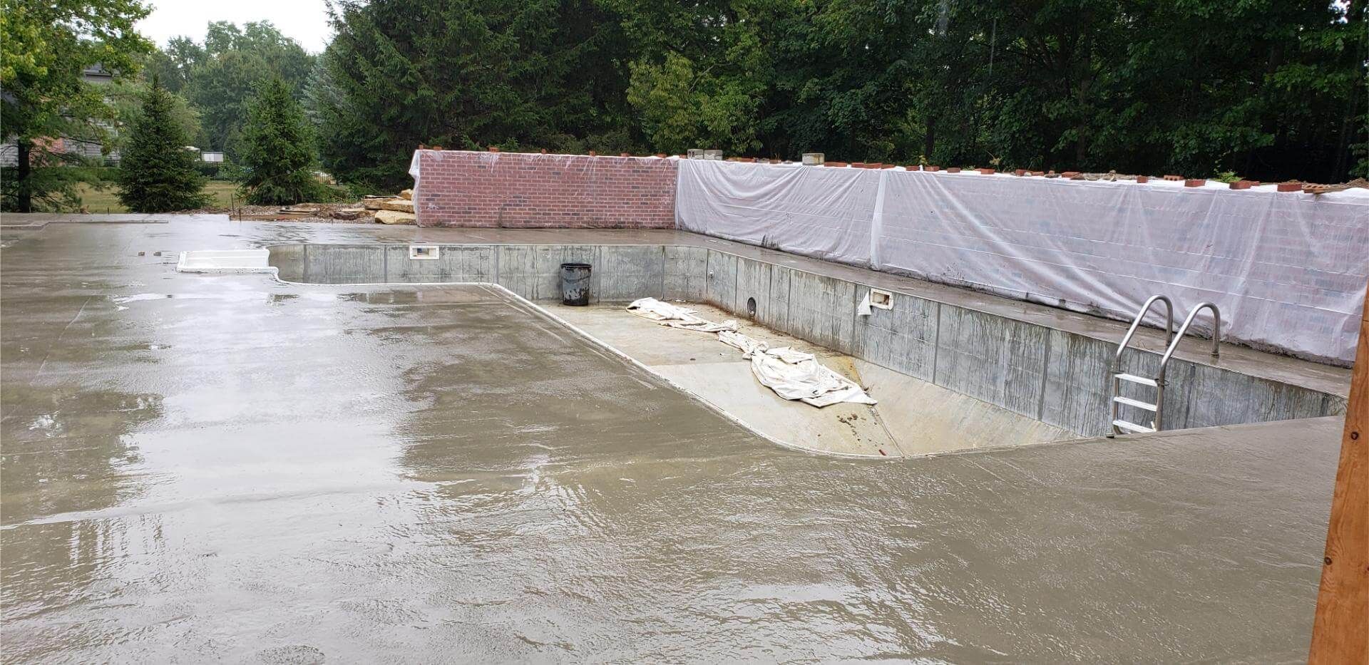 Concrete pool under construction, with a brick wall section and ladders, surrounded by wet concrete.