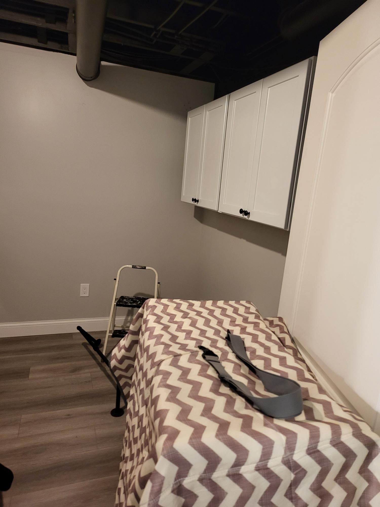 Gray wall, white cabinets, bed with chevron blanket, step ladder, and door in a room.