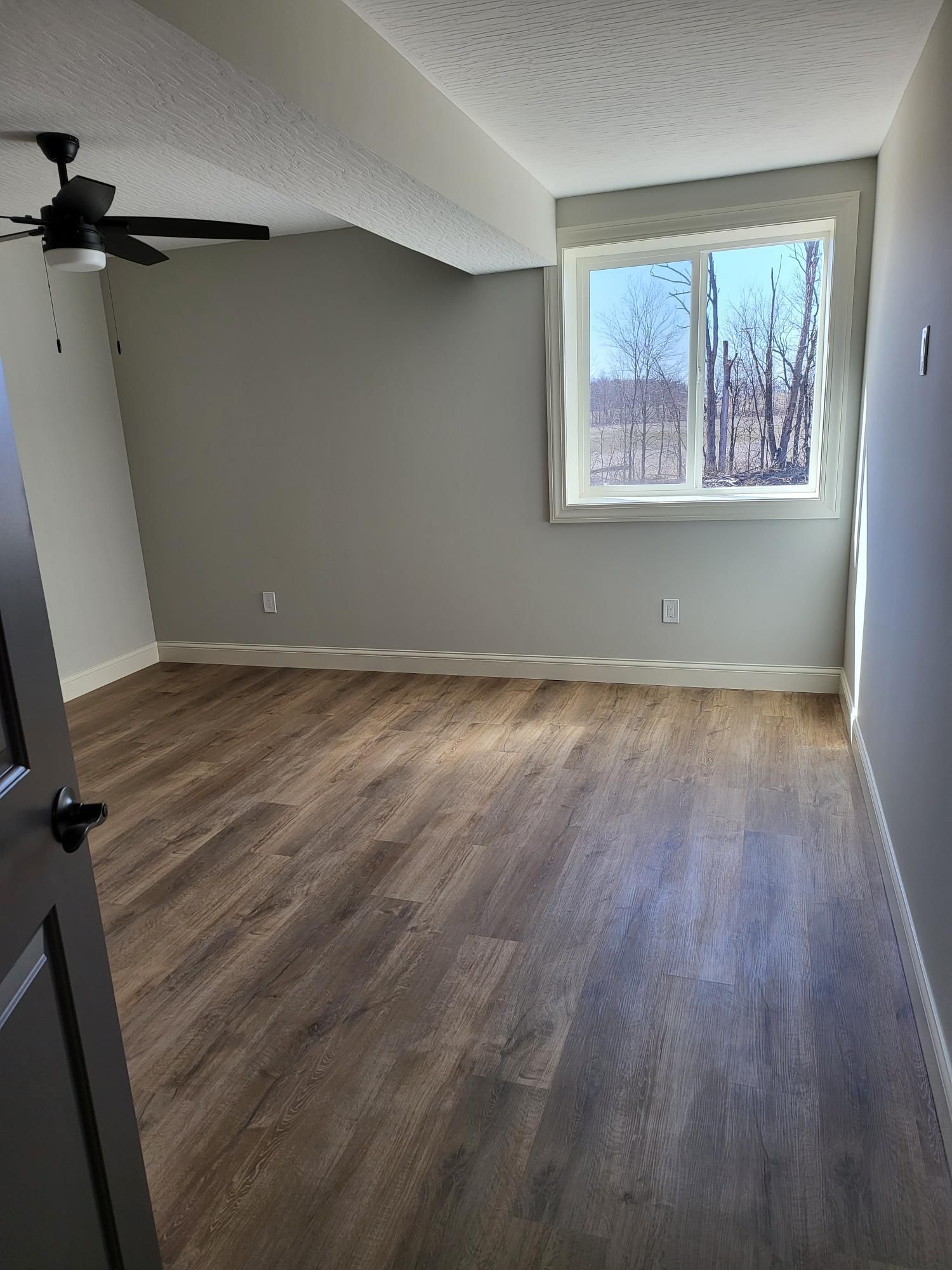 Empty bedroom with gray walls, brown floor, a window, and a ceiling fan.