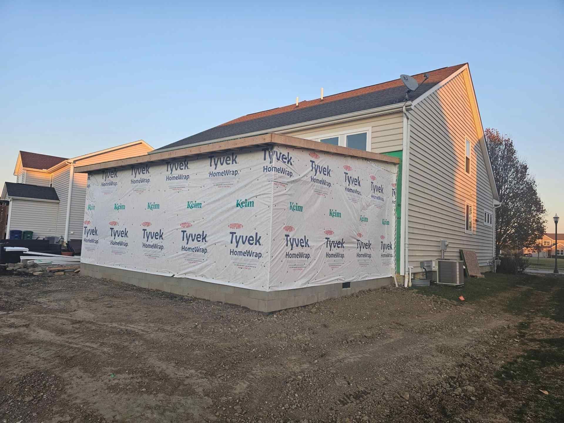 Exterior view of a house with a new addition under construction. Walls covered with Tyvek. Gravel yard.
