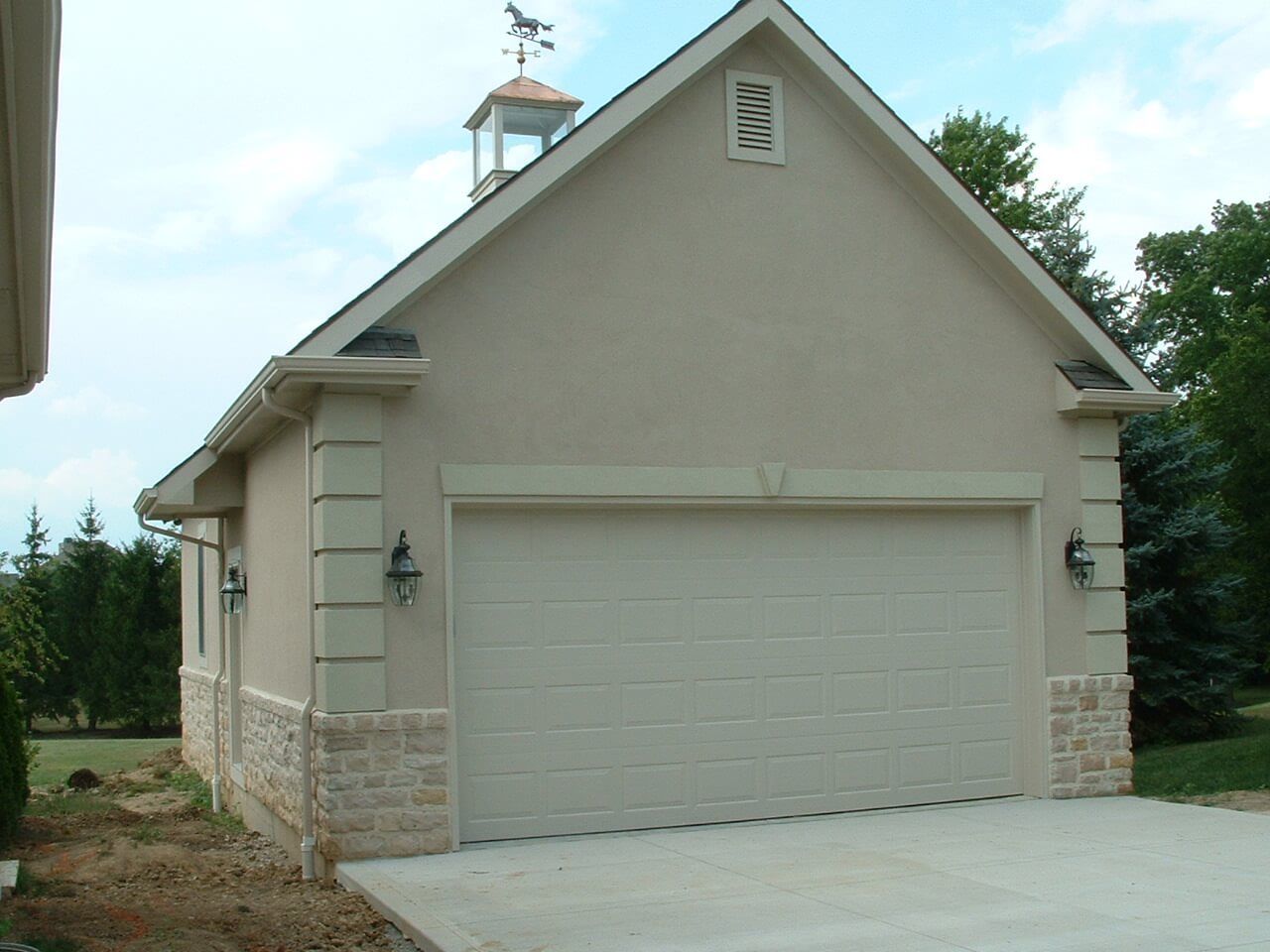 Tan stucco garage with stone accents, a weathervane, and a closed garage door.