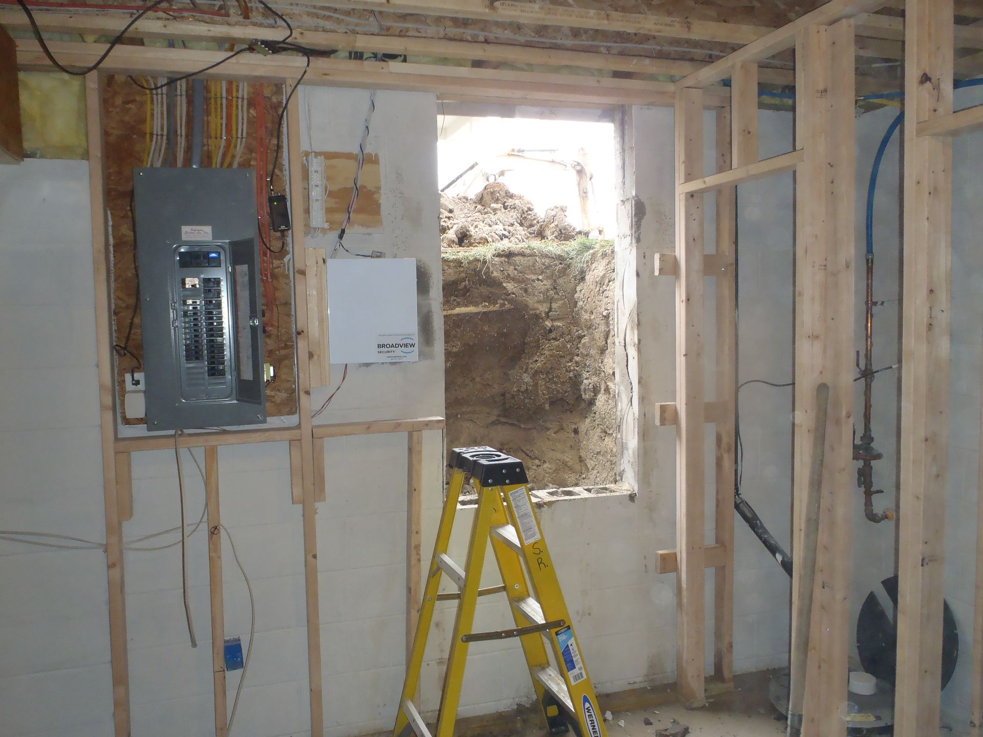 Basement interior with unfinished wooden framing, electrical panel, ladder, and a window looking out onto soil.