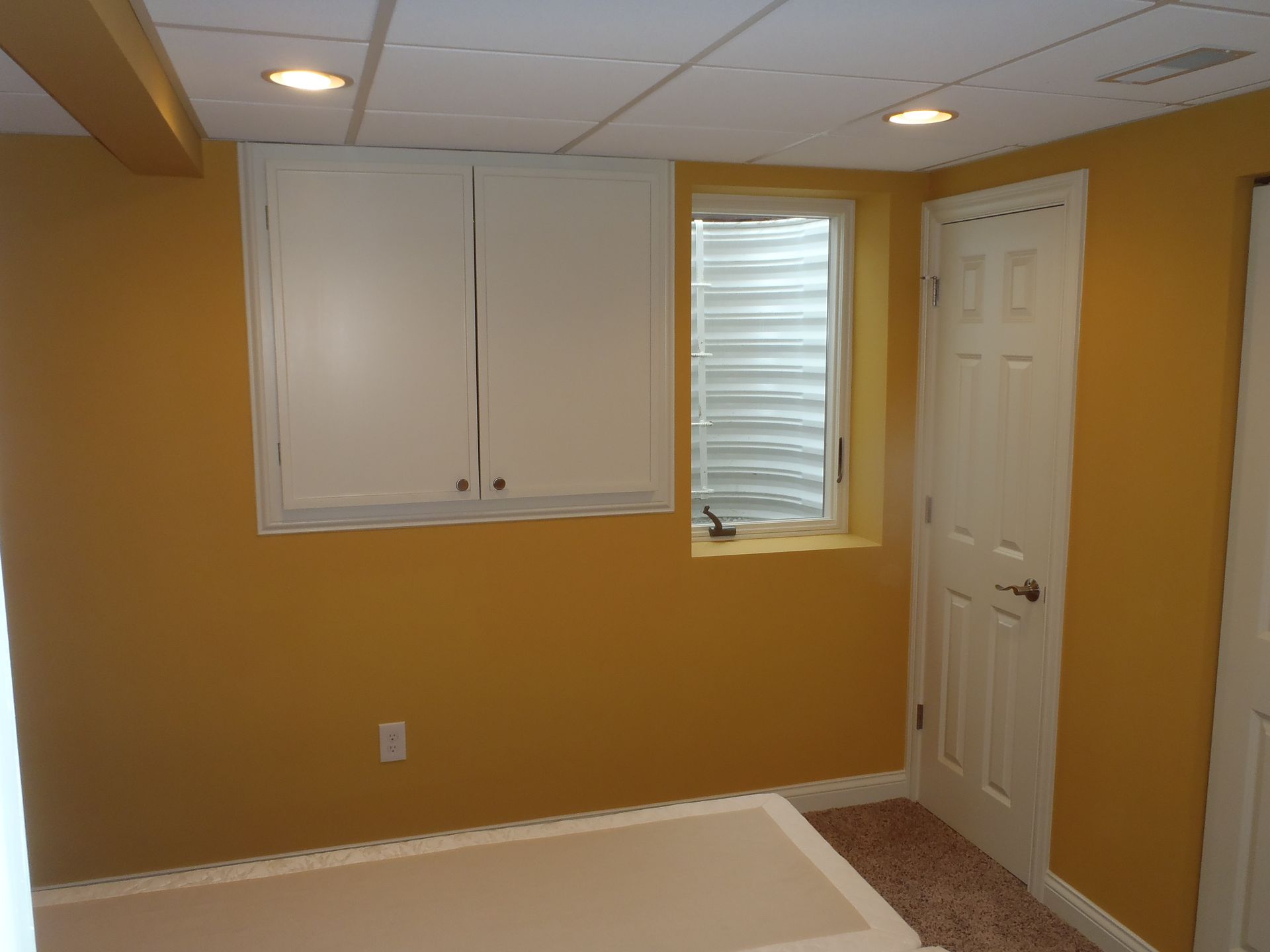 Yellow-walled basement room with white cabinets, window, and door; a cream-colored table sits at the bottom.