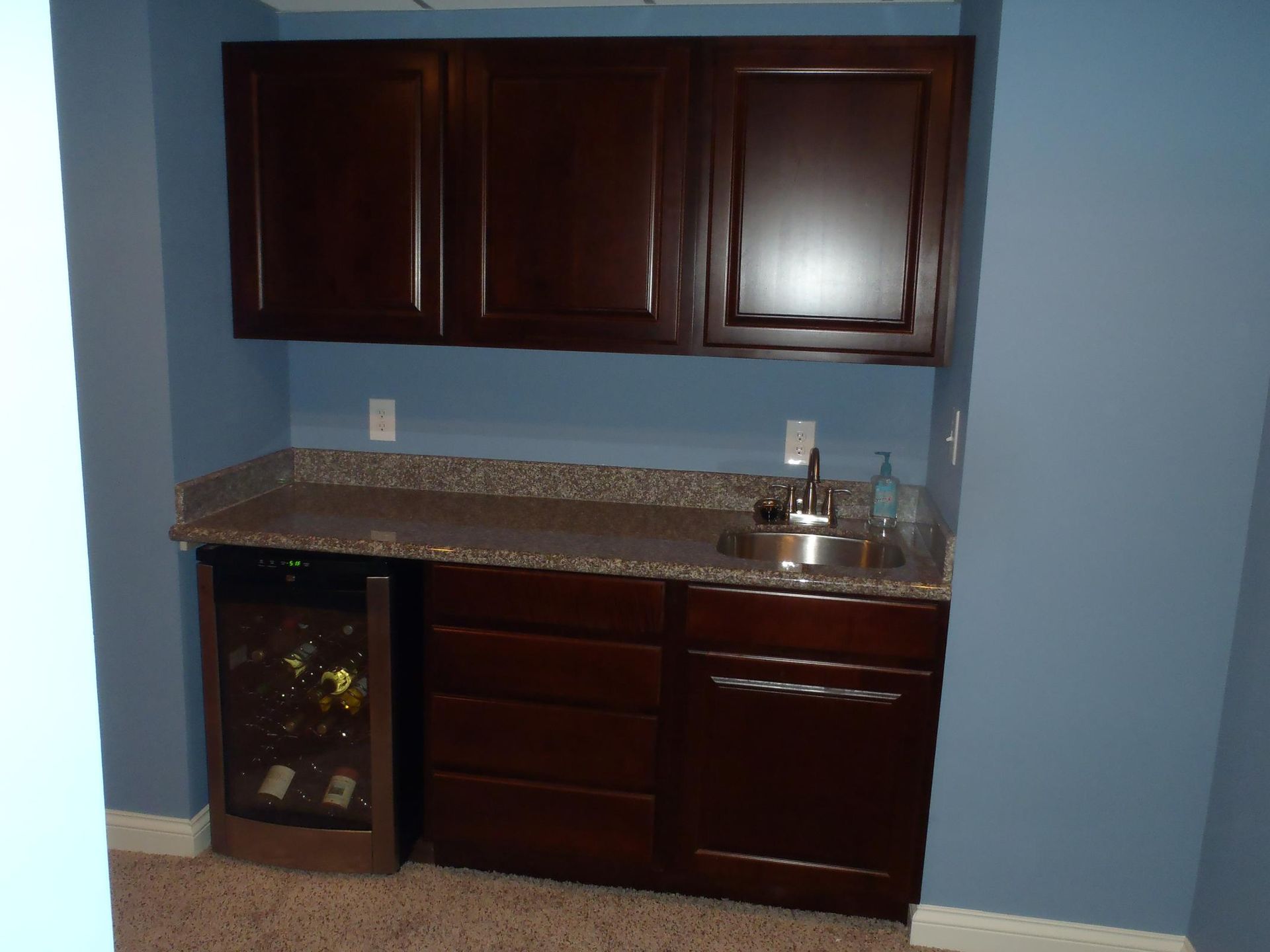 Dark wood wet bar with granite countertop, sink, and wine fridge in blue-walled room.