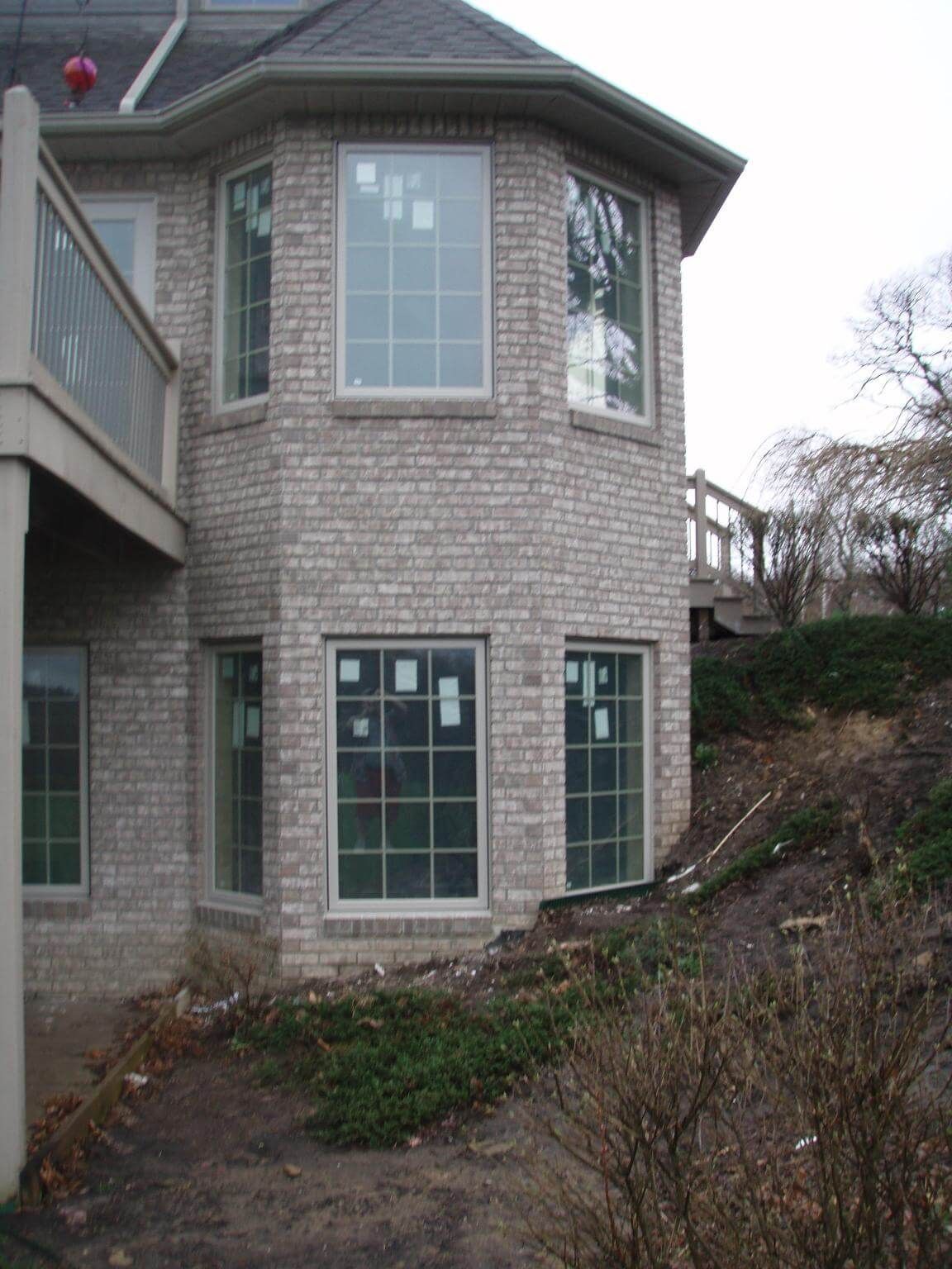 Brick house with bay windows, a deck, and a sloped yard with shrubbery.