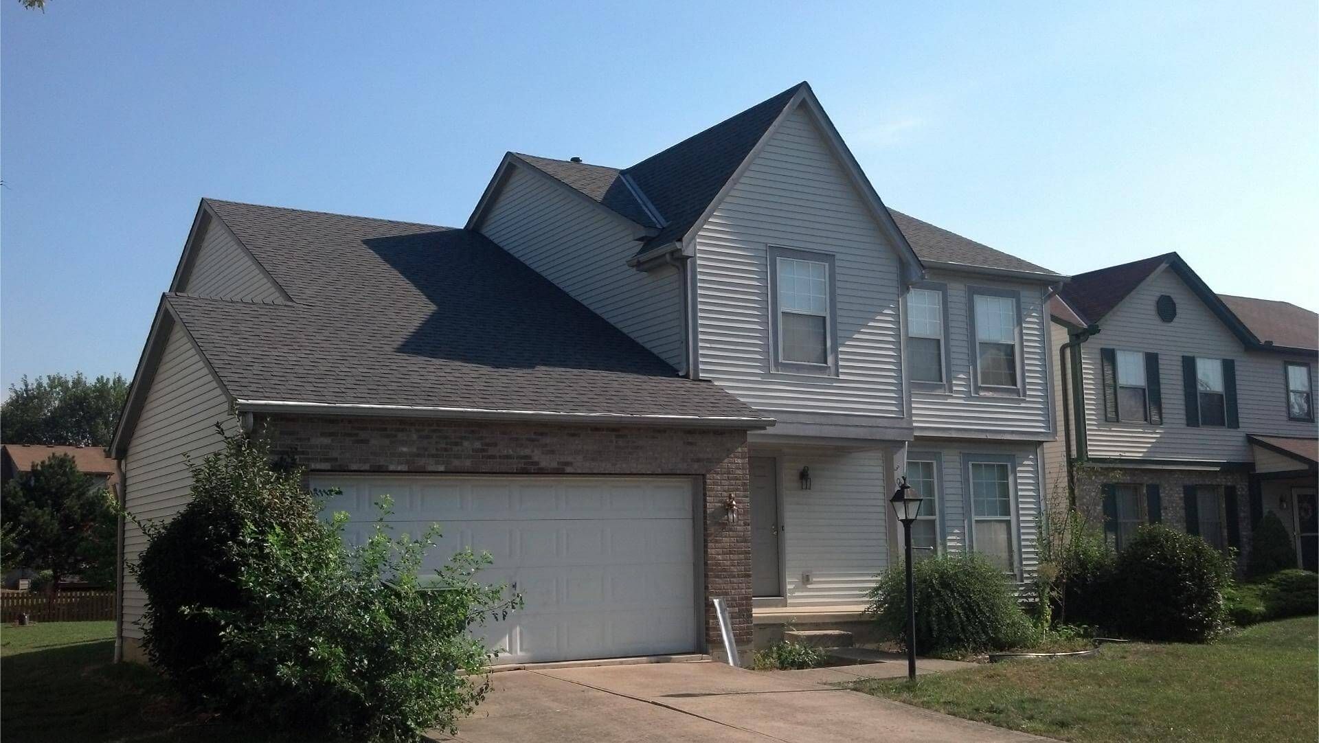 Two-story house with gray roof and white siding, fronted by a driveway and bushes, under a blue sky.