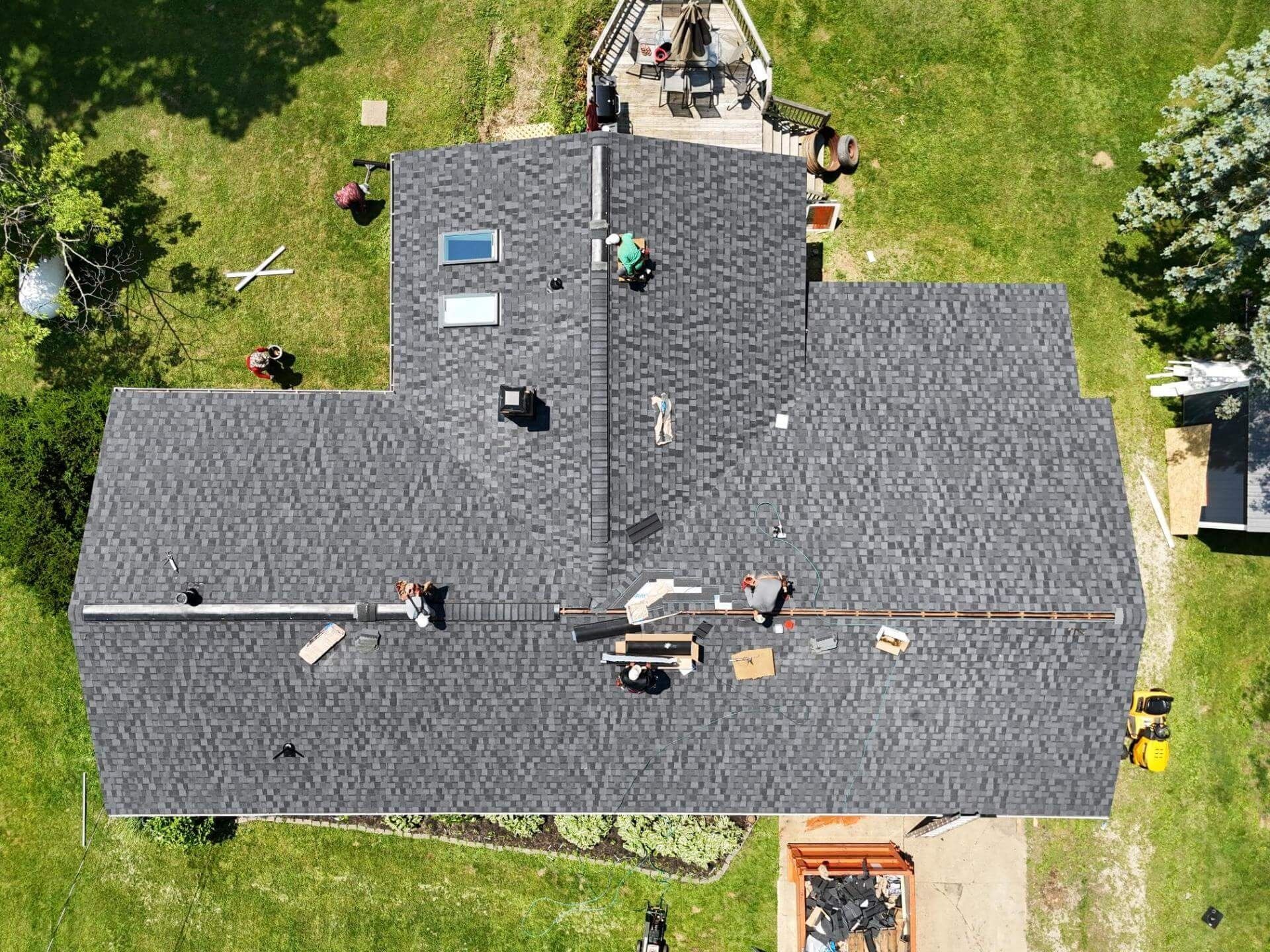 Overhead view of a house with roofers working on a dark gray shingled roof in a grassy yard.