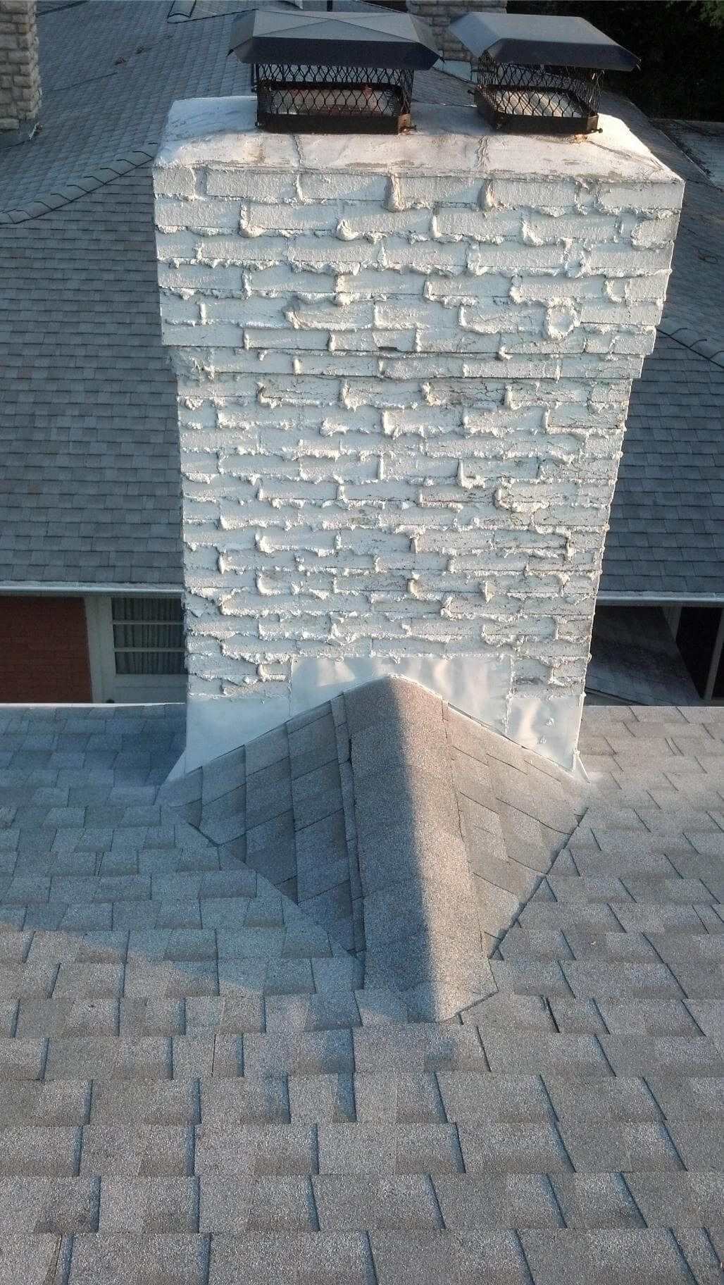 A chimney with peeling white paint, set against a gray shingle roof, has two metal caps on top.