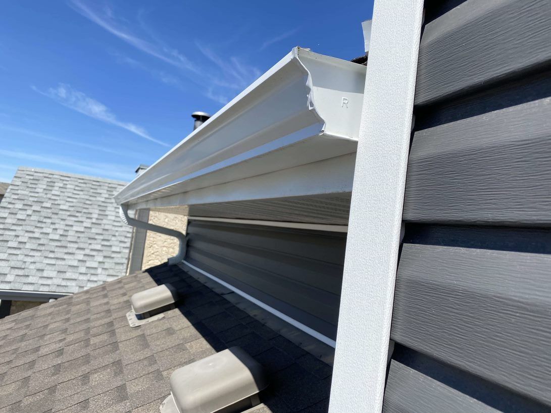 White gutter along the edge of a house with gray siding, a blue sky, and a glimpse of a gray rooftop.