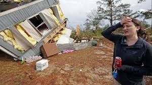 Woman stands distraught near a damaged mobile home after a disaster.