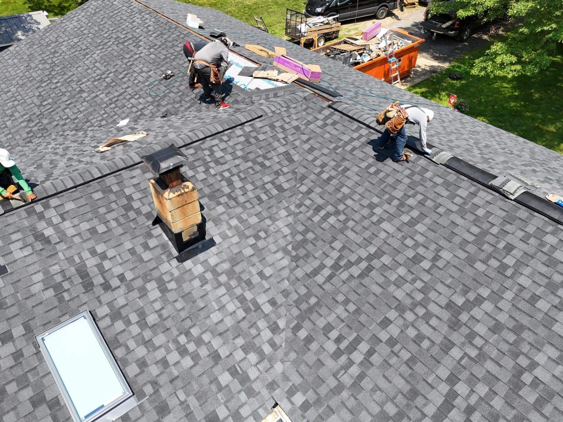 Roofers installing new shingles on a gray asphalt shingle roof, near a chimney and skylight.