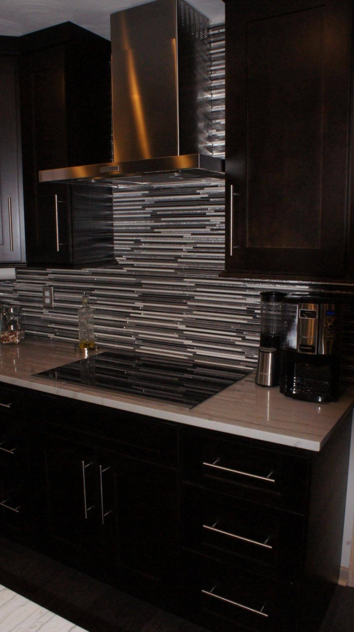 Dark kitchen with stainless steel hood, backsplash, cooktop, and cabinets.